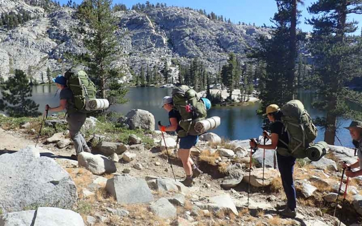 a group of people carrying backpacks hike past a body of water with mountains in the background